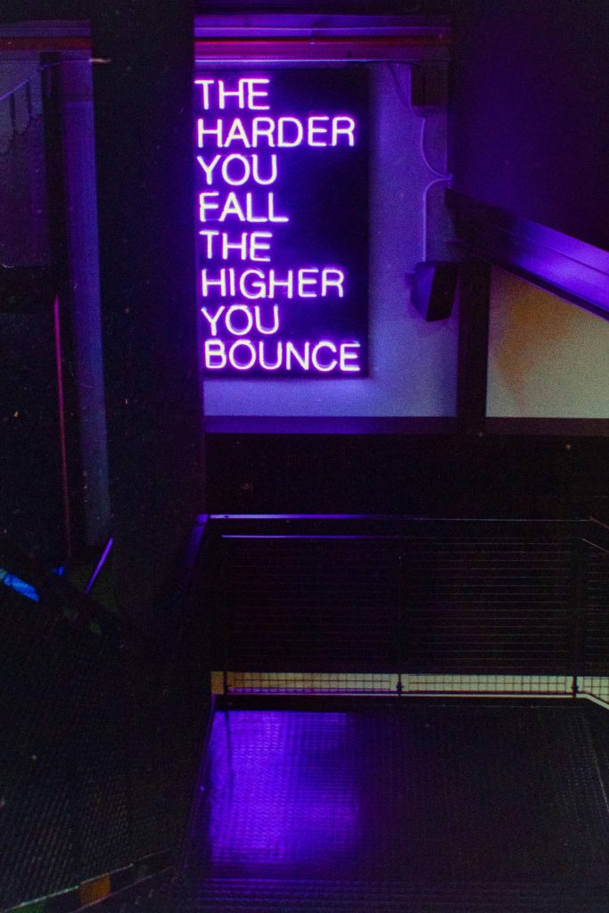 Una scala illuminata da luci blu e viola con un’insegna al neon che recita ‘THE HARDER YOU FALL THE HIGHER YOU BOUNCE’.