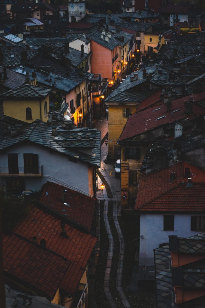 Vista aerea di una strada tortuosa in un antico borgo al crepuscolo. Gli edifici sono strettamente affiancati e mostrano una varietà di colori caldi, con luci gialle che illuminano il percorso e alcune finestre. La scena evoca un’atmosfera accogliente e intima, con il contrasto tra la luce naturale che svanisce nel cielo e le luci artificiali