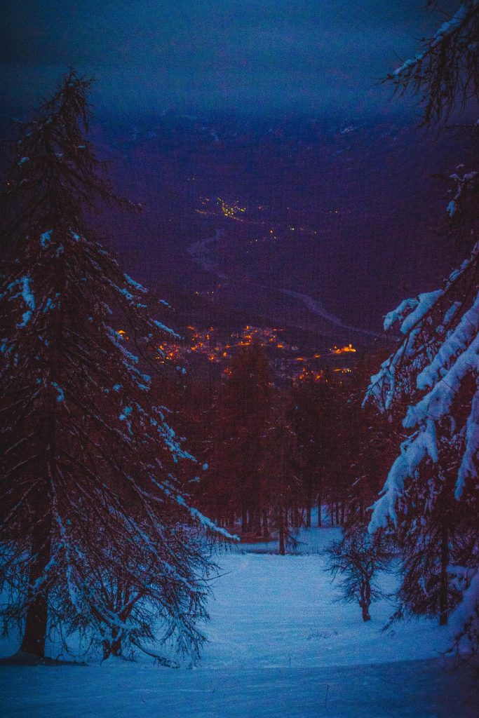 Una vista serale con una città illuminata in lontananza, in primo piano dei pini innevati fanno lateralmente da cornice e in basso possiamo vedere un esteso prato innevato, La luce è crepuscolare e misteriosa e suggerisce che la foto è stata scattata appena dopo il tramonto