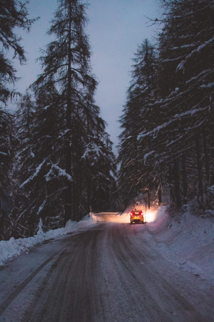 Strada innevata fiancheggiata da pini carichi di neve durante il crepuscolo o la prima serata. Il cielo è crepuscolare, suggerendo che il momento della giornata è appena dopo il tramonto. Un veicolo con le luci posteriori accese è visibile in lontananza, che si allontana dal punto di vista.