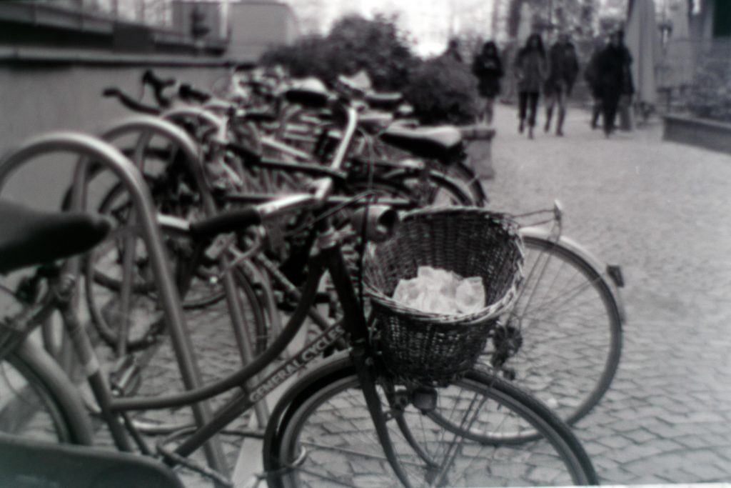 Una rastrelliera per biciclette piena di biciclette parcheggiate. sullo sfondo si vedono delle persone e si distingue il pavimento in ciottoli di pietra