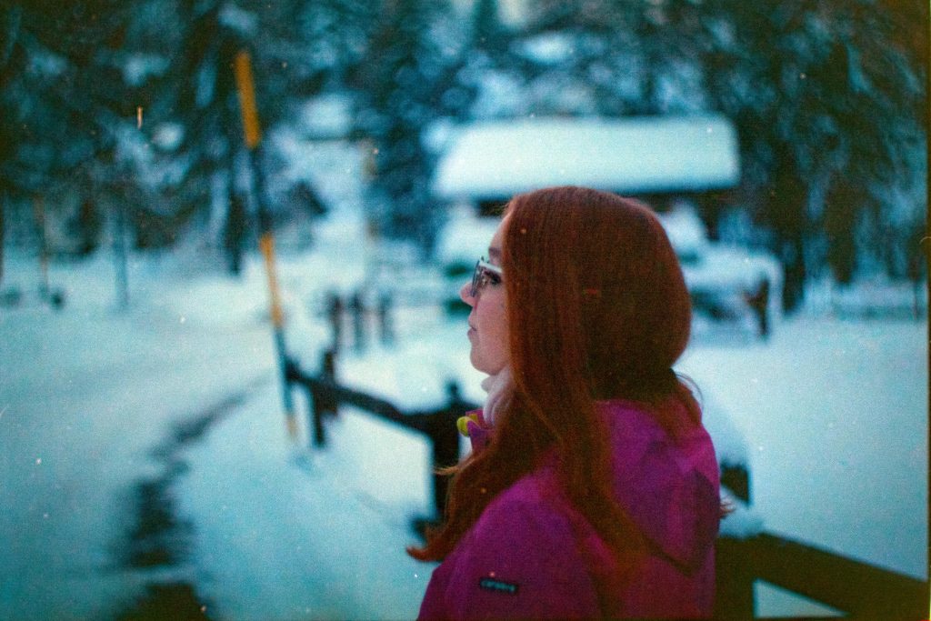 Una persona dai capelli rossi osserva contemplativa il paesaggio innevato, avvolta in una giacca viola. Sullo sfondo, una scena invernale sfocata di alberi e neve, che dona un'atmosfera tranquilla e riflessiva