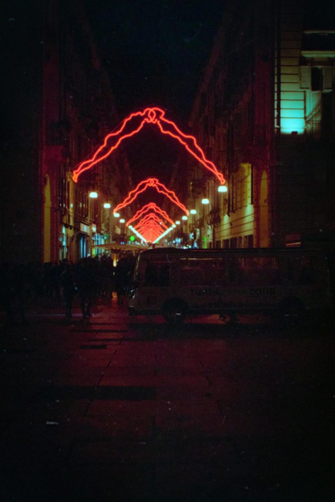 Una via illuminata da luci di natale a forma di persone che si baciano si estende in lontananza e illumina una piccola folla dietro ad un bus del trasporto pubblico