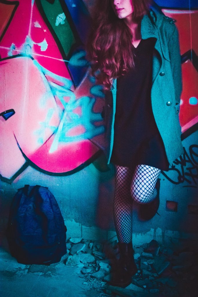 A girl standing in front of a graffiti in a run down place, the eyes are not seen and the colours are altered in order to give the photo a surreal atmosphere, the girl's hair are slightly pink, the graffiti is bright pink and everything around is turquoise