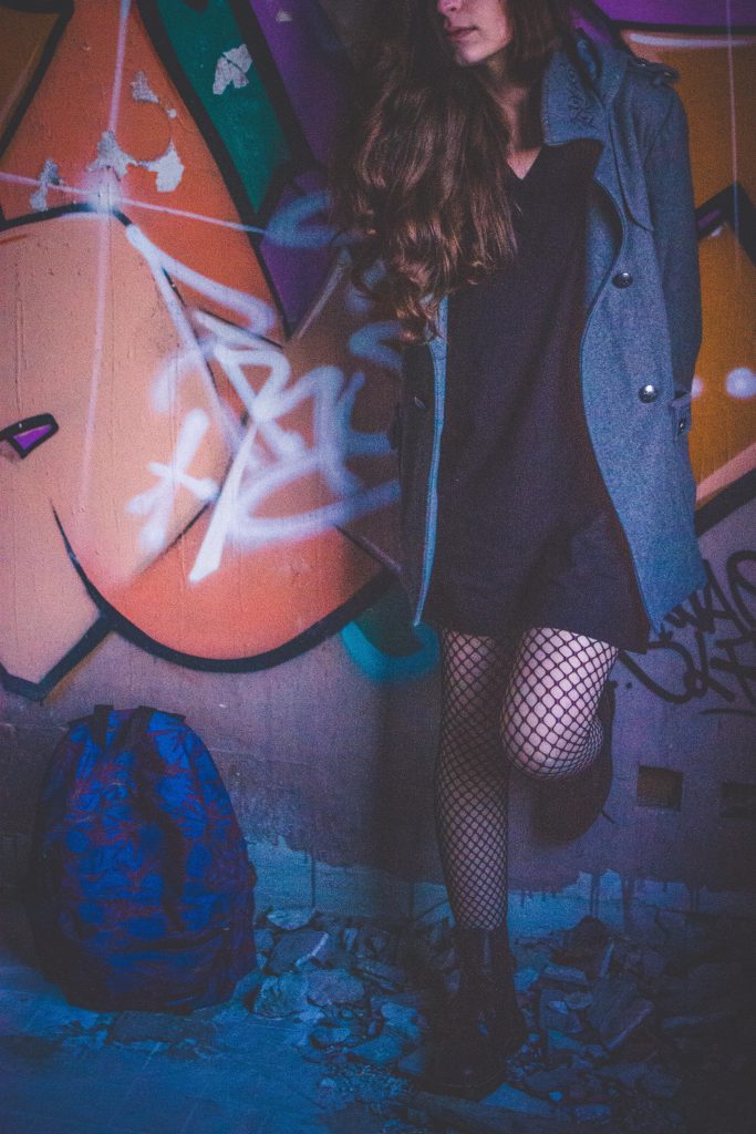 A girl standing in front of a orange graffiti in a run down place, the eyes are not seen