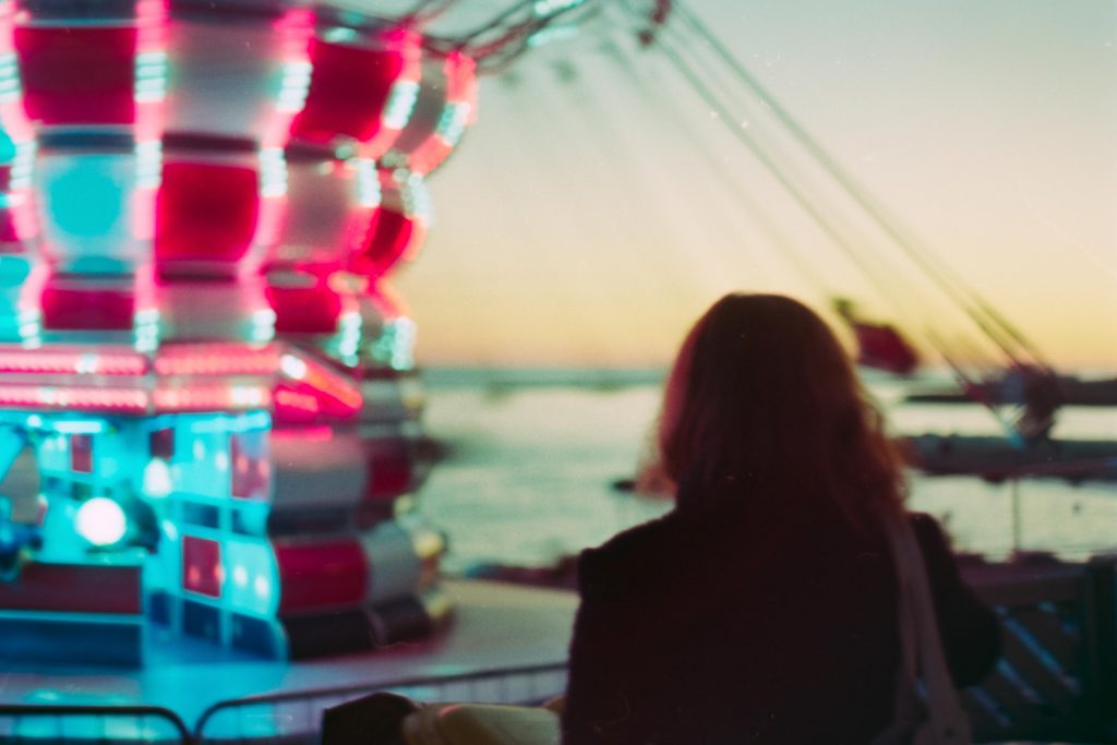 A girl viewed from the back is looking at a carousel lit by pink and blue lights. In the background we can also see the sky at sunset