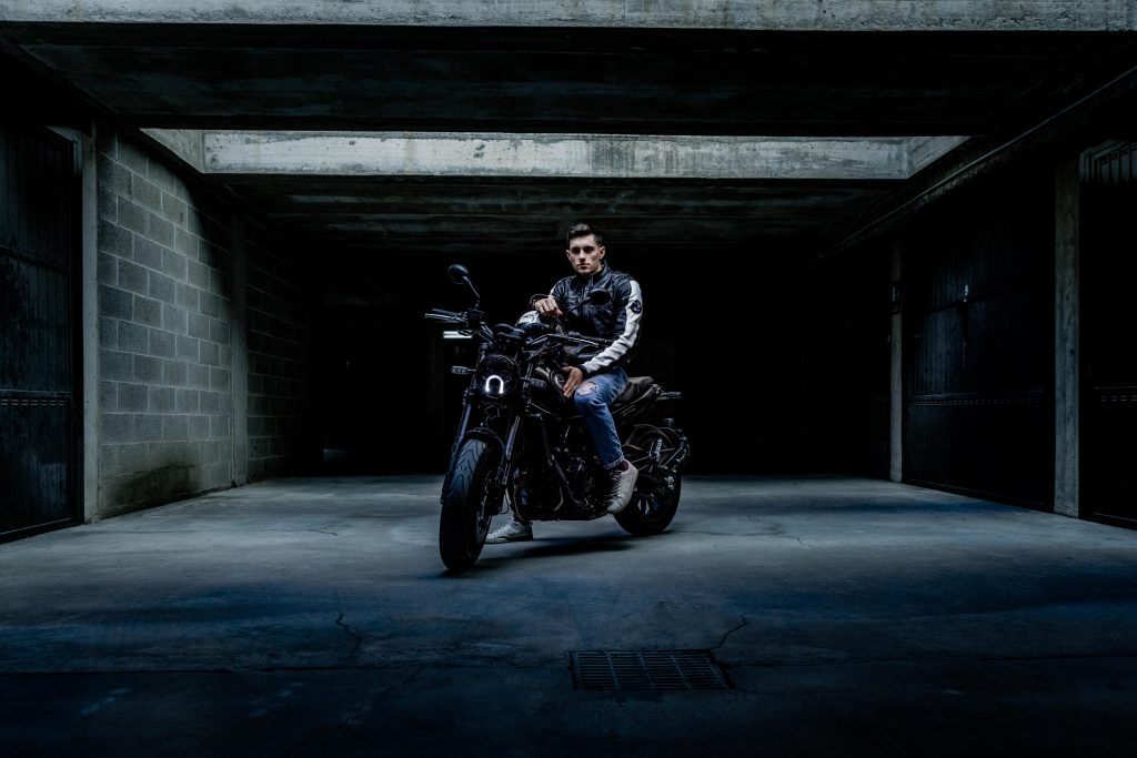 A guy wearing jeans and a leather jacket sitting on a parked bike in an underground parking lot, seen from the front