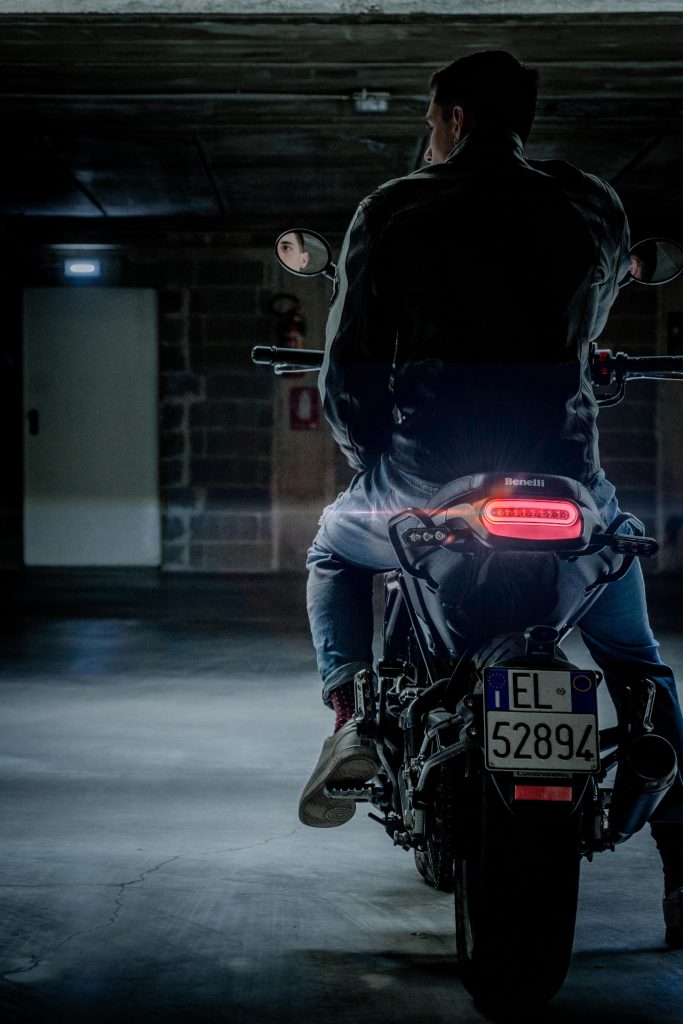 A guy wearing jeans and a leather jacket sitting on a motorcycle with lights on, inside an underground parking spot