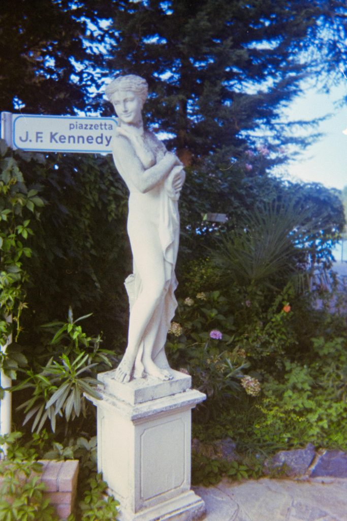 a fake granite statue representing a woman in front of a fake street sign with "viale j.k. Kennedy" written on it