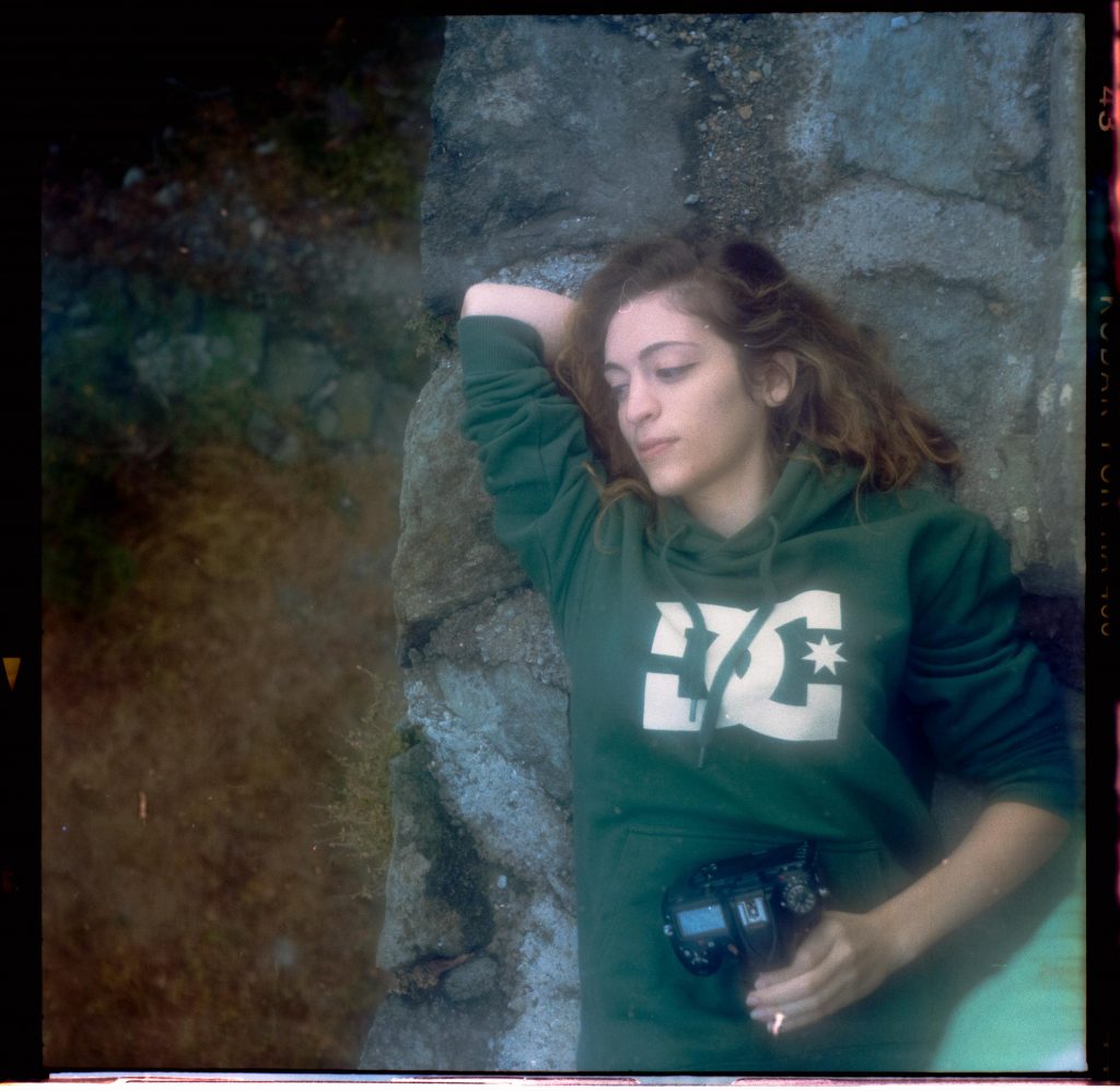 A girl with a green hoodie resting on a stone construction with a camera in his hands