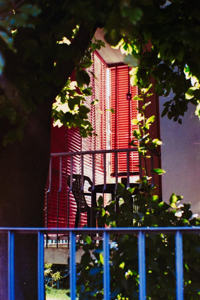 A balcony with a dark green plastic table and chairs, lit by the sunlight filtering from a red venetian curtain, in front of this scene we can see greenery, a tree and a blue fence in the very front