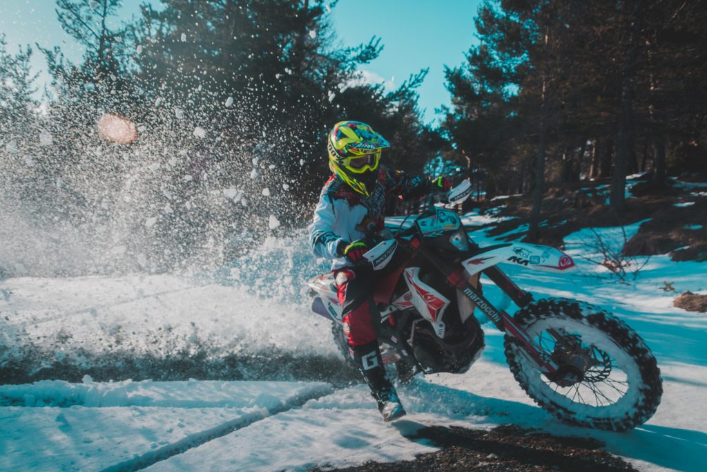 A rider with red and white motorcycle clothes and a white dirt bike drifting in the woods on a patch of snow, kicking up snow with the rear wheel