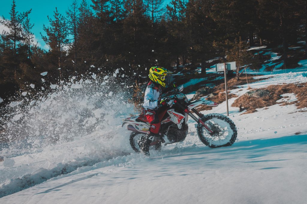 A rider with red and white motorcycle clothes and a white dirt bike drifting in the woods on a patch of snow, kicking up snow with the rear wheel