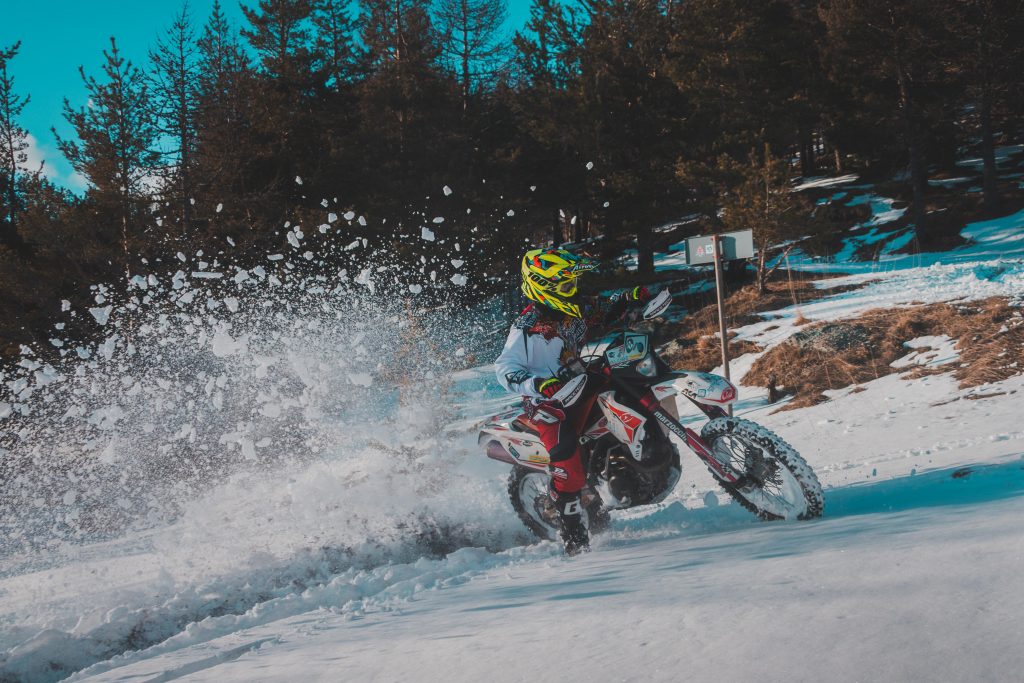 A rider with red and white motorcycle clothes and a white dirt bike drifting in the woods on a patch of snow, kicking up snow with the rear wheel