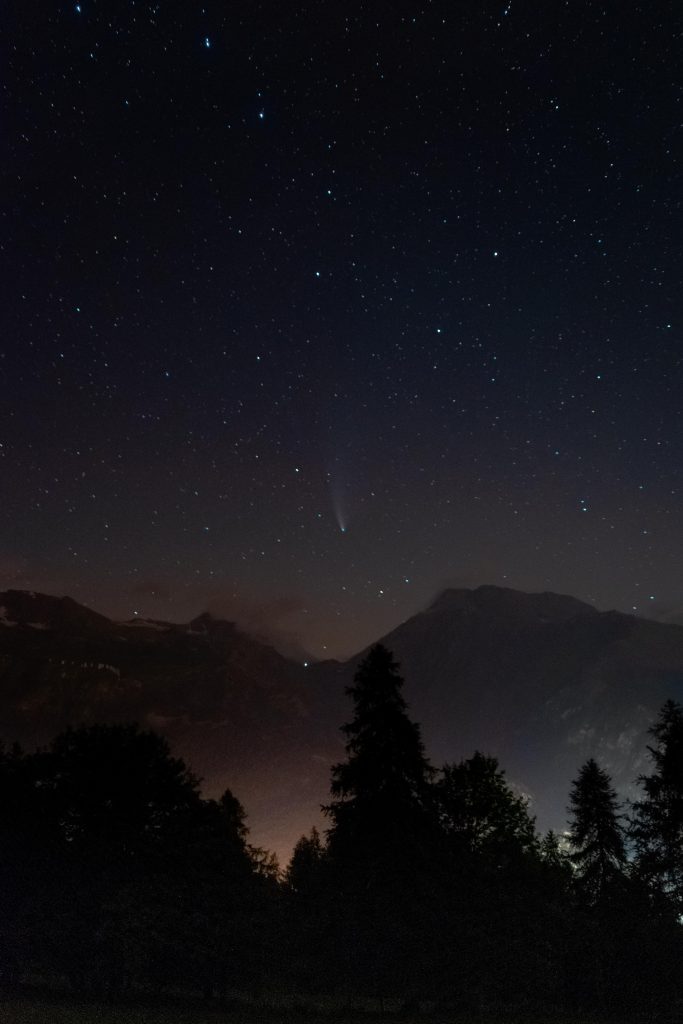 A mountain night landscape with light pollution on the bot, tree silouhettes in the front, mountains in the back and a starry night on top, in the center, a comet seen from a distance with it's trail