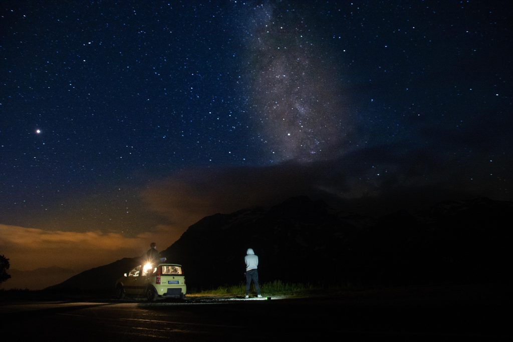 un ragazzo seduto sul tetto di una macchina ferma al bordo di una strada e un altro ragazzo con il cappuccio in piedi affianco alla macchina, di fronte a loro un panorama montano con sopra delle nuvole e il cielo stellato con la Via Lattea visibile