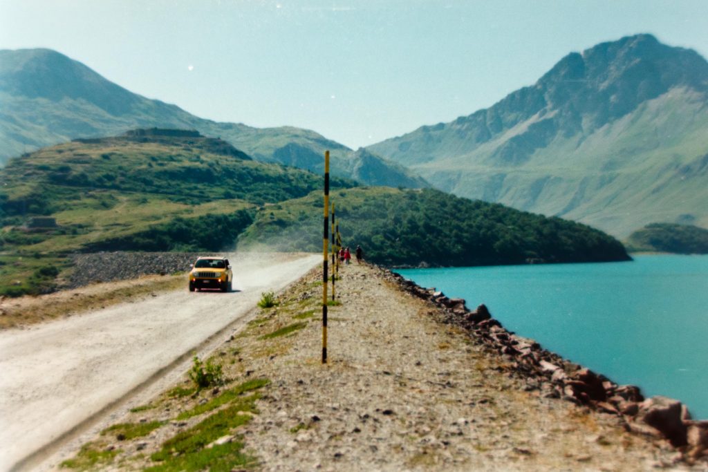 A yellow jeep travelling through a dirt road with black and yellow snow poles on the road side and a lake on the back with mountains on the background