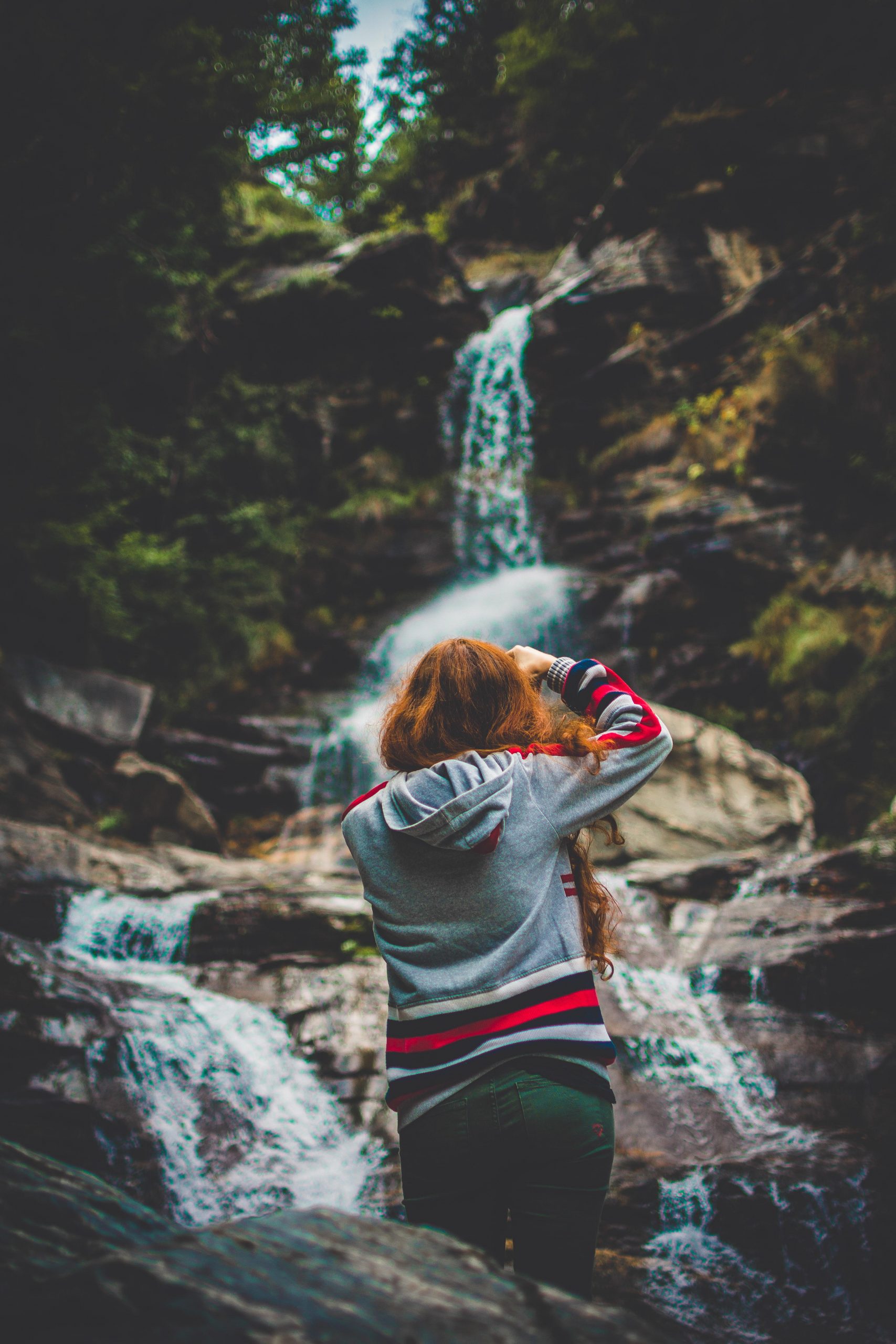 Una ragazza con i capelli rossi e mossi con addosso una maglia grigia che scatta una foto ad una cascata