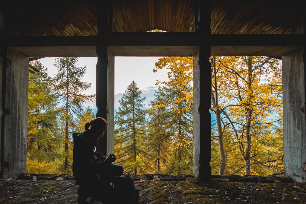 a man crouching to reach his backpack on the floor in front of a big broken window with no more glass and trees outside