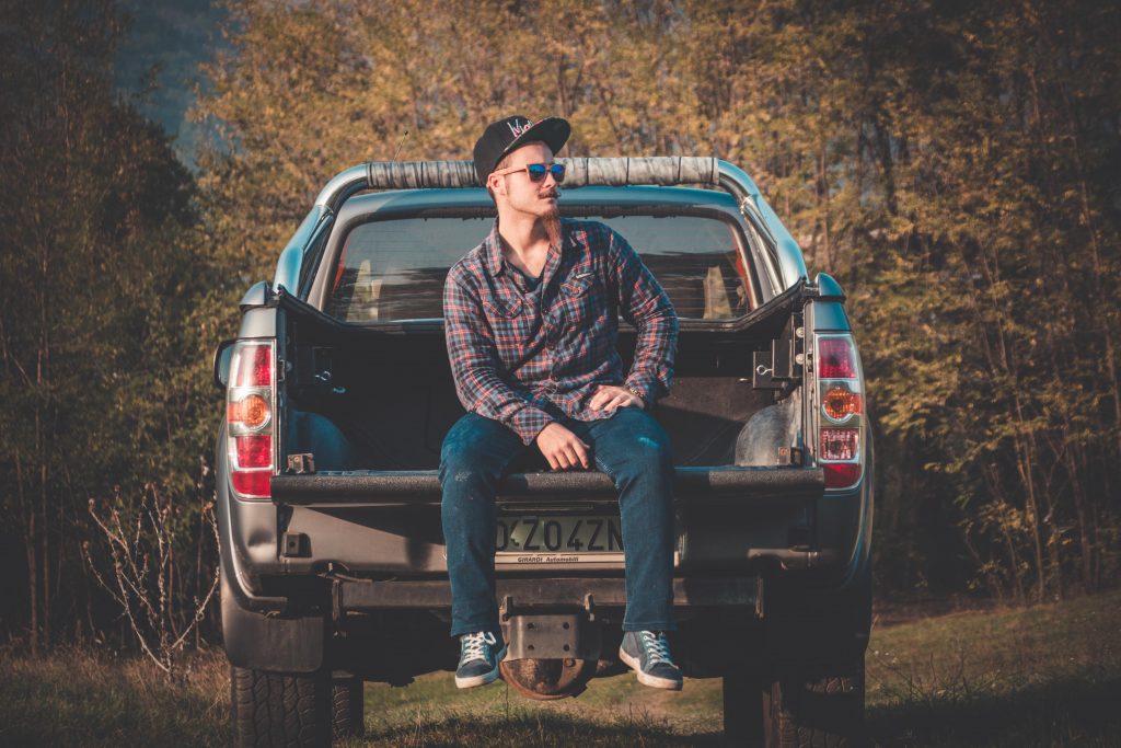 Guy with a flanel and a long beard is sitting on a pick-up truck's bed