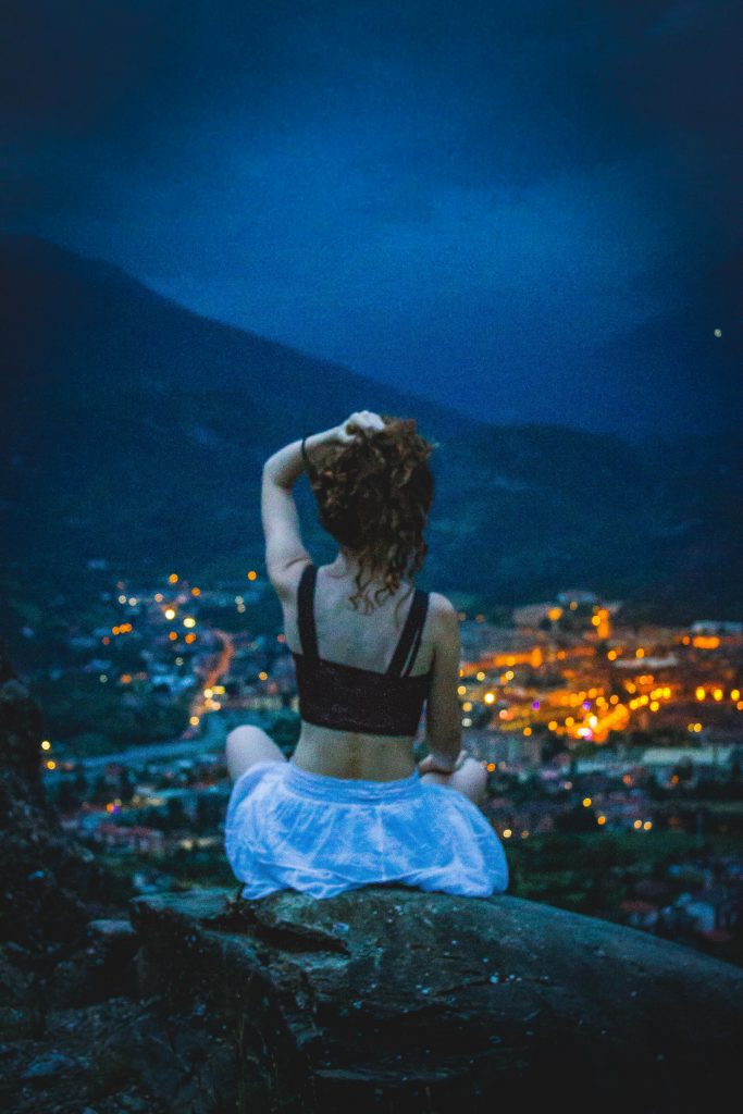 A girl in a white skirt and a black top sitting on a rock overlooking a lit up city