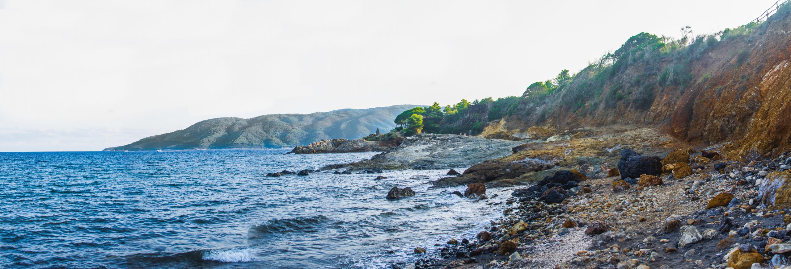 A big panorama picture of a wild Beach of Elba Island with black sand, light brown rocks, the sea on the left, a hill on the right and on top of the hill there is run down old minerary equipment and trees around it